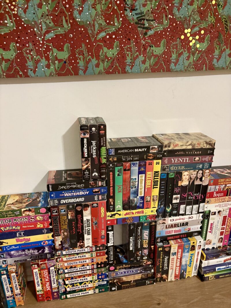 A large assortment of VHS tapes is stacked against a wall beneath a colorful abstract painting. The tapes include movies, TV shows, and cartoons, with titles visible on their spines.