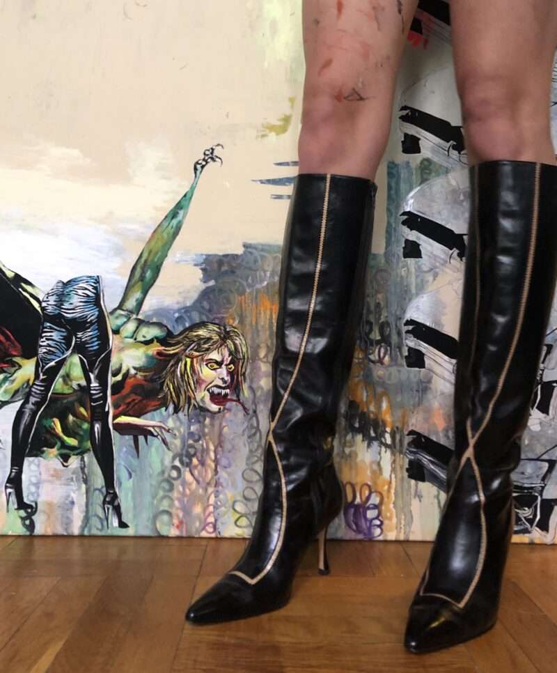 Person wearing black heeled boots stands in front of a painting featuring a monstrous, humanoid creature with exaggerated features and an aggressive expression.