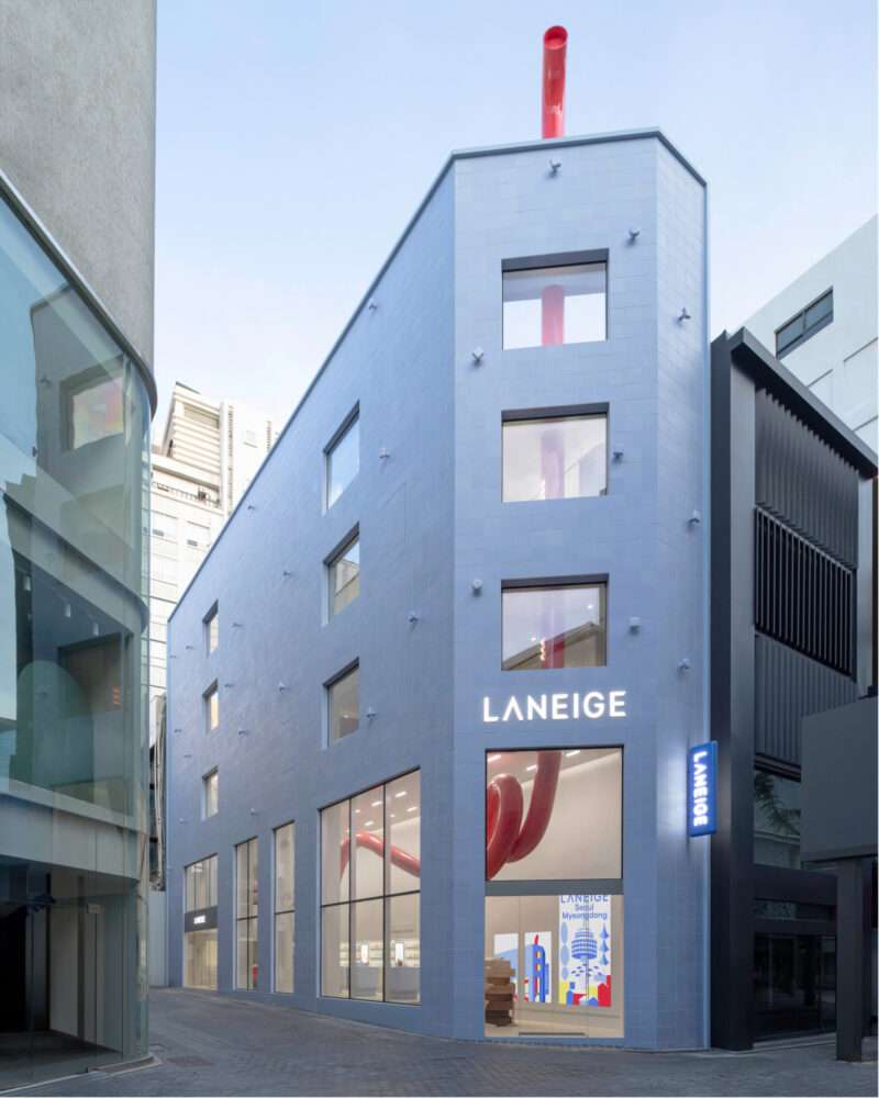 A modern, light blue building with large windows and “LANEIGE” signage stands on a city street, showcasing the Laneige Seoul flagship with a prominent red structure visible inside.