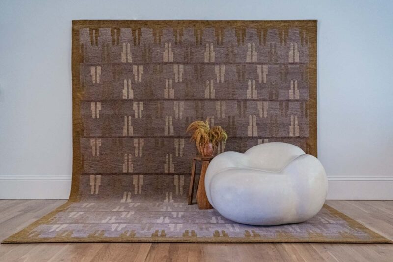 A modern white chair and small wooden table with dried flowers sit on a patterned brown rug that extends up the wall, against a plain background and wooden floor.
