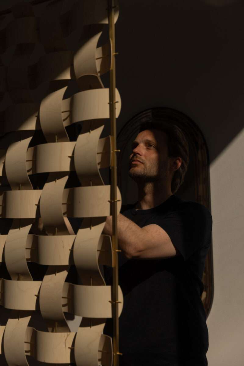 A man in a dark shirt stands next to a wooden art installation with curved pieces, illuminated by a beam of light in an otherwise shadowed room—a scene reminiscent of Rick Tegelaar’s innovative approach to material and design.