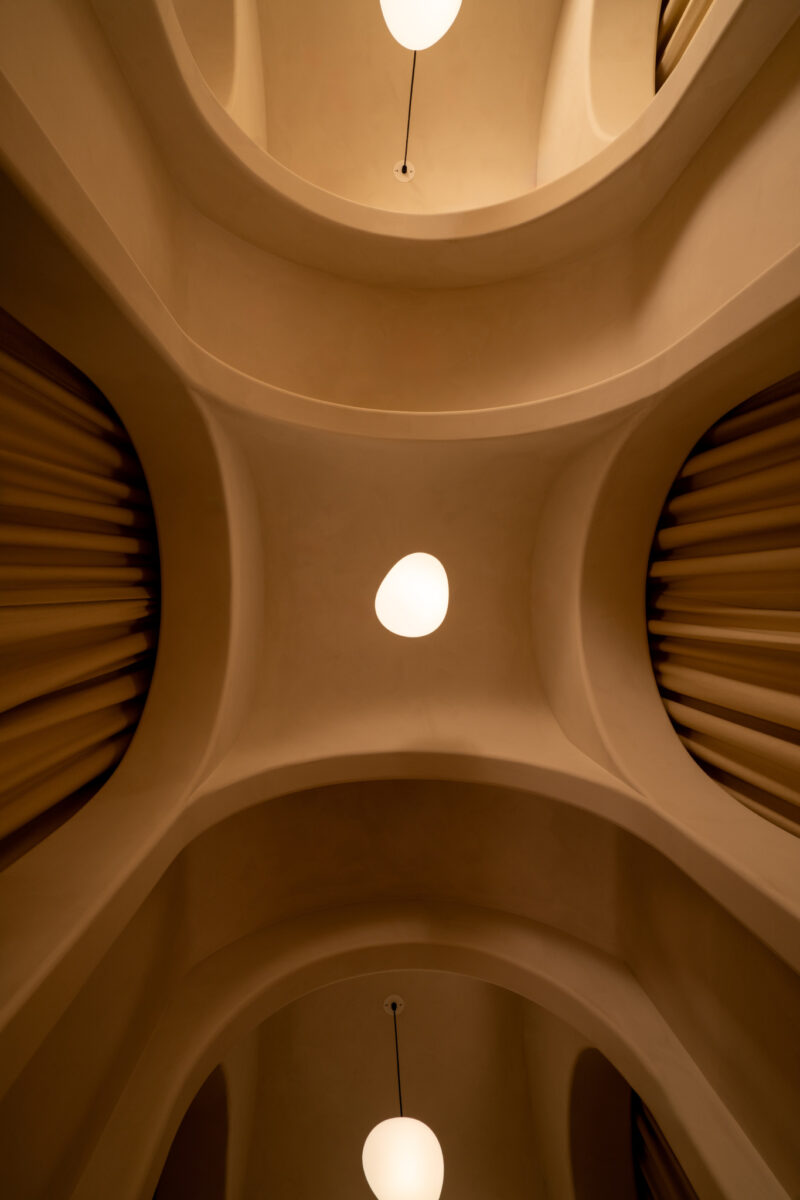 Curved architectural ceiling at Sana Skin Studio Wynwood features three hanging round lights and ribbed wall details, viewed from below. Warm, beige color tones dominate the scene.