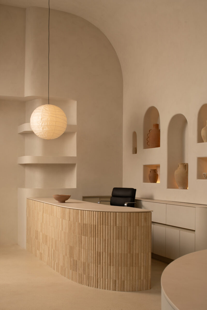 Curved wooden reception desk with a black chair, built-in shelves, and pottery accents in a beige minimalist room at Sana Skin Studio Wynwood, beautifully lit by a round pendant lamp.