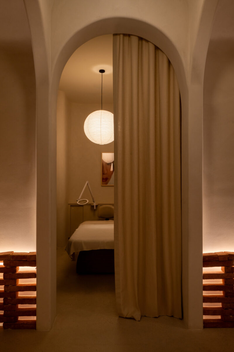 A softly lit treatment room at Sana Skin Studio Wynwood features a massage table, a round paper lantern, a curtain partition, and minimalist decor in neutral tones.