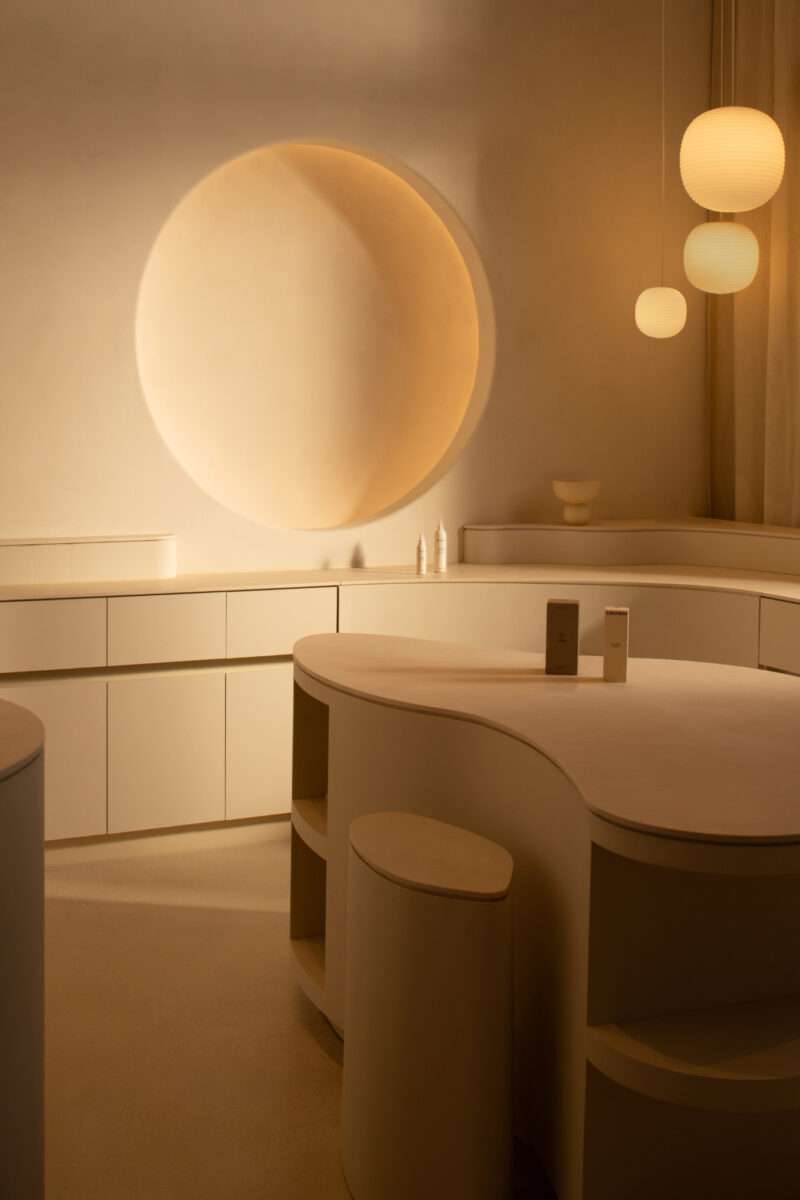 Minimalist beige room with curved counters, round wall feature, three hanging globe lights, and two bottles on the counter—capturing the serene elegance of Sana Skin Studio Wynwood.