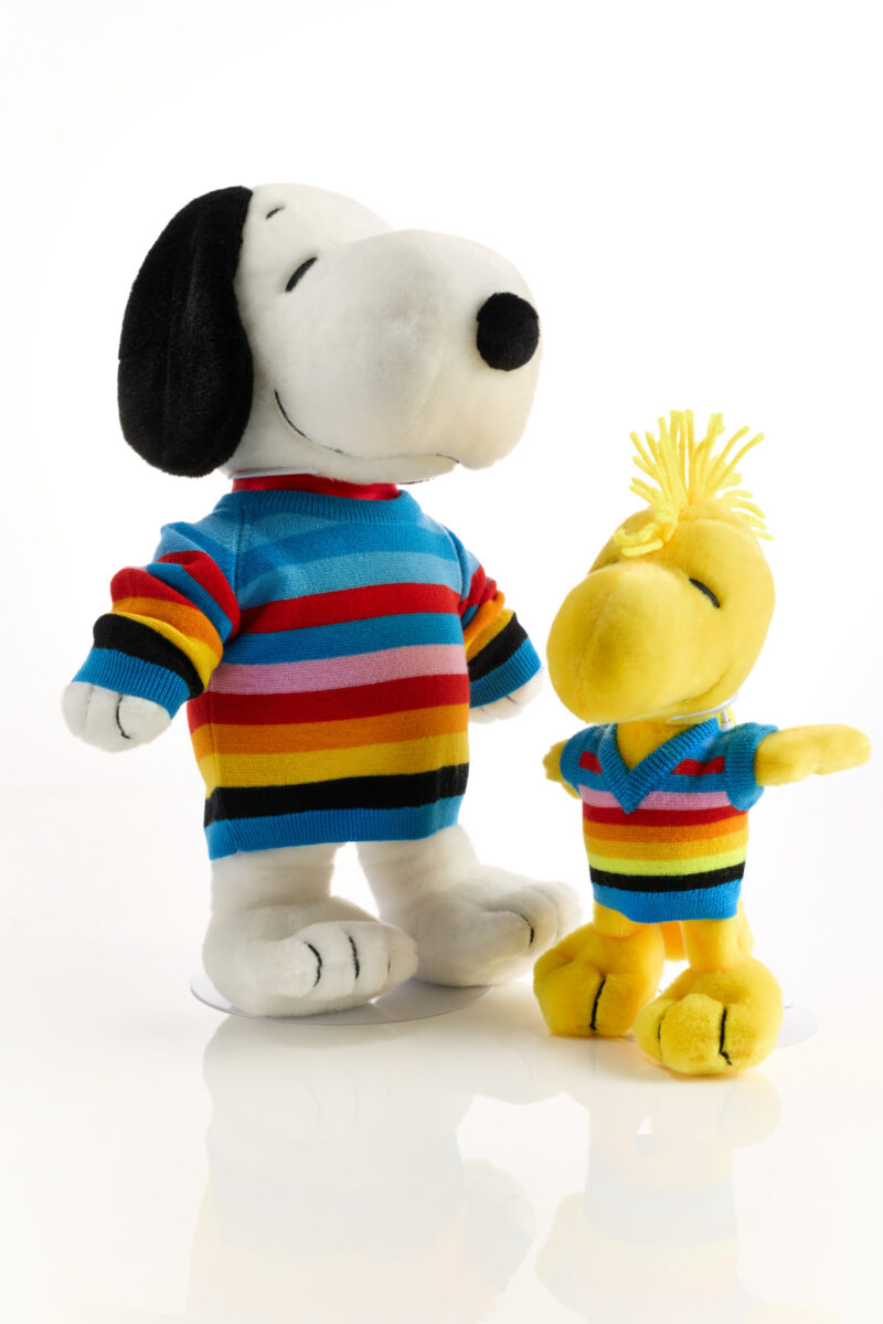 Two plush toys, a black-and-white dog and a yellow bird, both wearing matching rainbow-striped sweaters, are posed against a white background—bringing playful Snoopy in Style NYC vibes to any space.