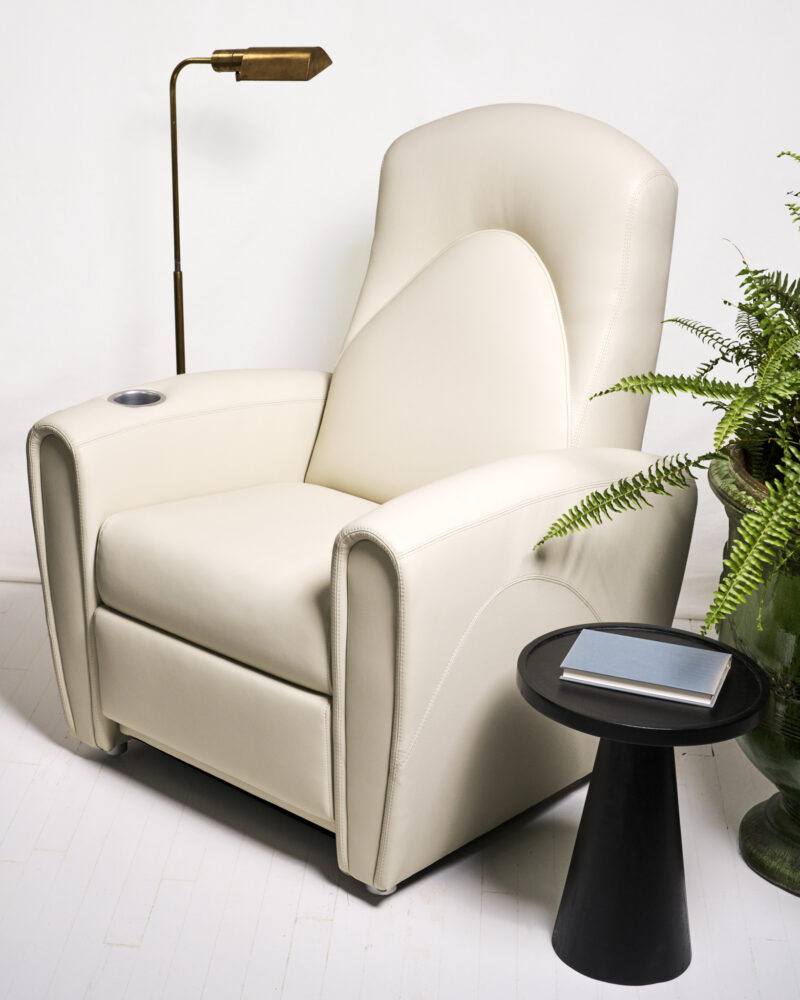 Beige recliner chair next to a small black side table with a closed book, a potted fern, and a brass floor lamp, all set on a white floor and background.