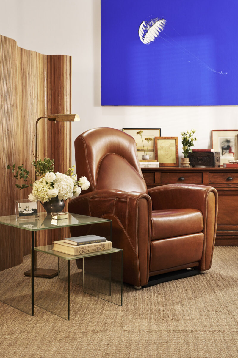 A brown leather armchair sits next to a glass side table with books and flowers, in a cozy room with wooden furniture, art, and framed photos.