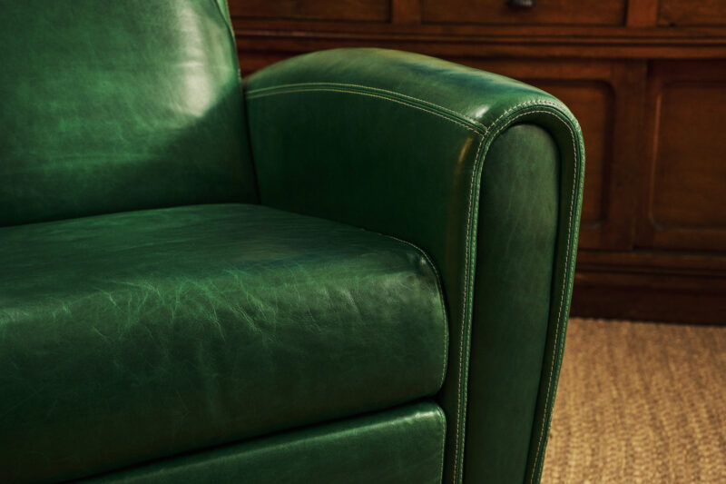 Close-up of a green leather armchair with visible stitching, set on a textured beige carpet with a wooden cabinet in the background.