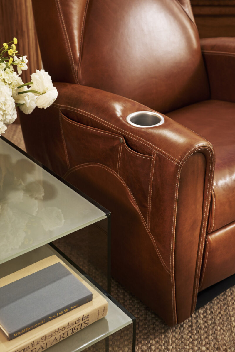 Brown leather armchair with a built-in cup holder, next to a glass-top table holding books and a vase of white flowers.