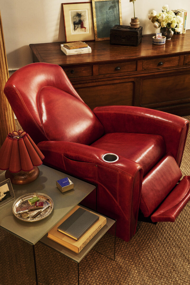 Red leather AWEchair luxury lift chair beside a glass side table with books and a tray, set in front of a wooden cabinet with framed art and flowers.