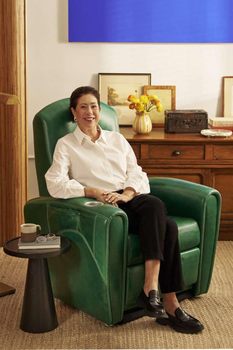 A woman in a white blouse and black pants sits smiling in a green armchair next to a small table with a mug, glasses, and a book, in a warmly decorated room.