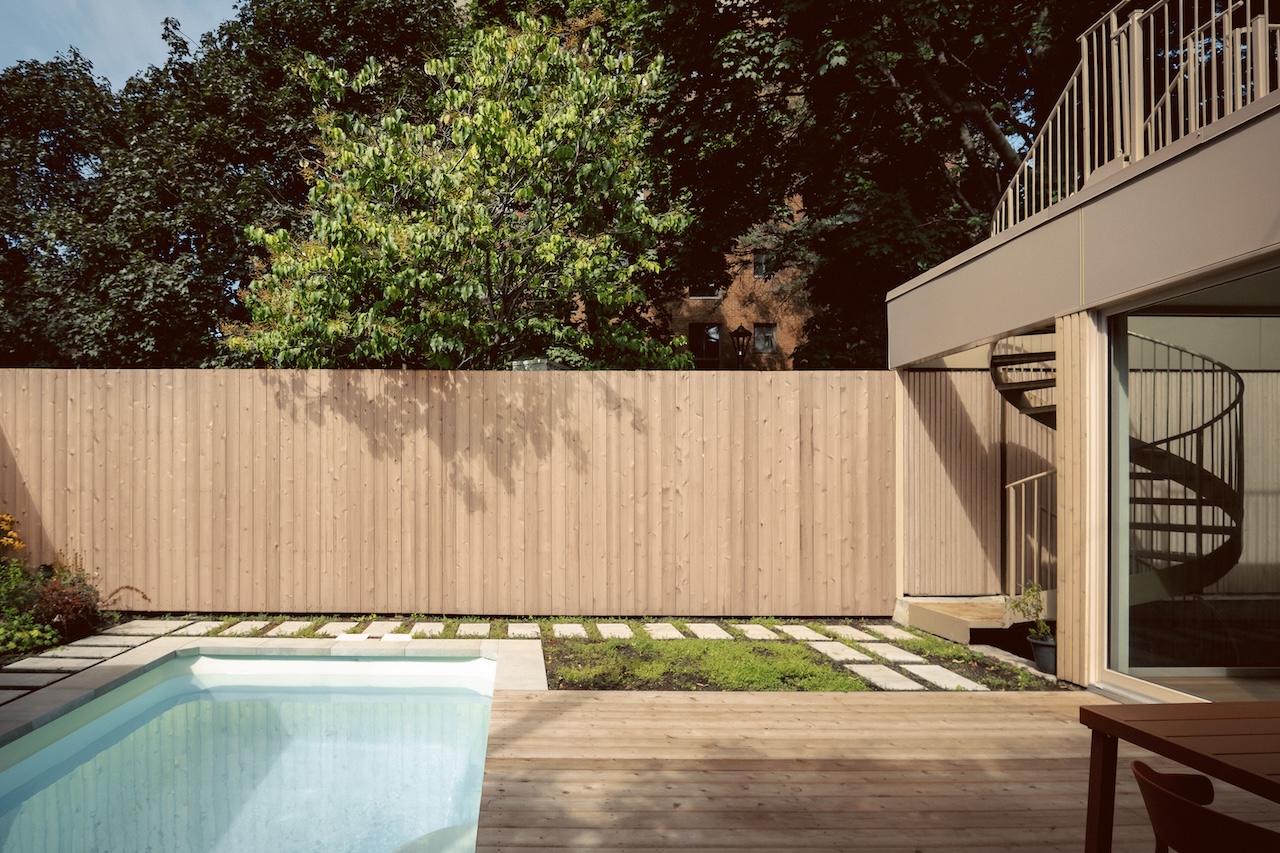 Modern backyard with a small pool, wooden deck, stone path, tall wooden fence, trees in the background, and a metal spiral staircase leading to an upper level.