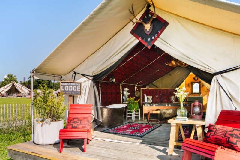 Glamping tent with open flap shows rustic interior, red chairs on porch, potted plant, and decorative rug, with a “Lodge” sign and antlers above entry. This inviting Urban Cowboy Tent sits amid grass with other tents visible in the background.