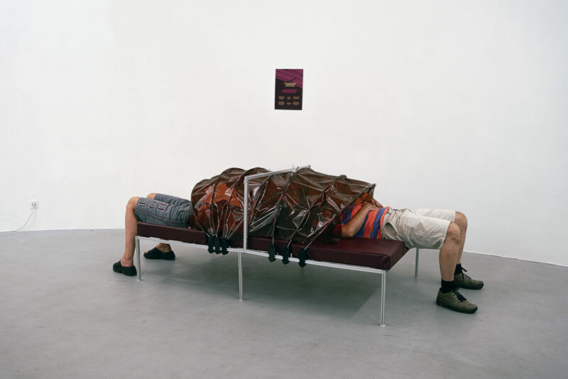 Two people lie at opposite ends of a bench, their upper bodies covered by a brown plastic shroud, with a small poster on the wall behind them.