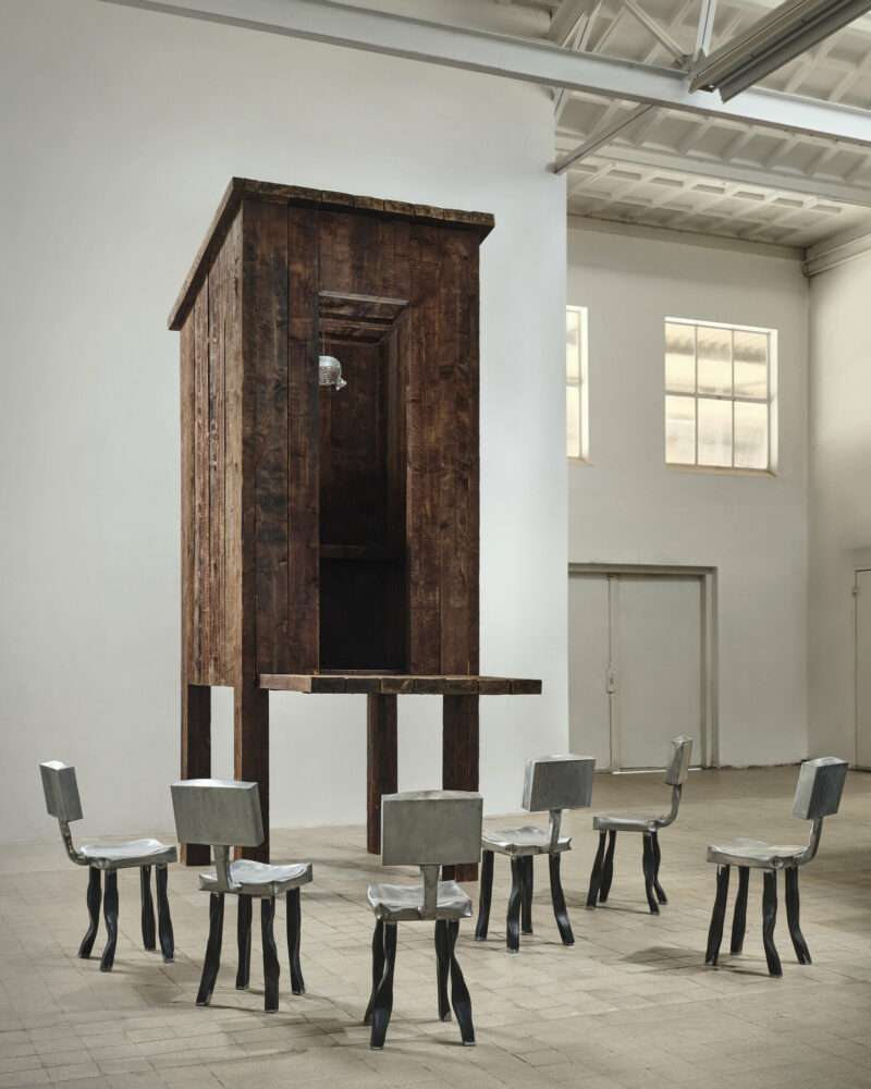 A tall, wooden structure resembling an outdoor privy on stilts, surrounded by six metallic chairs arranged in a circle.