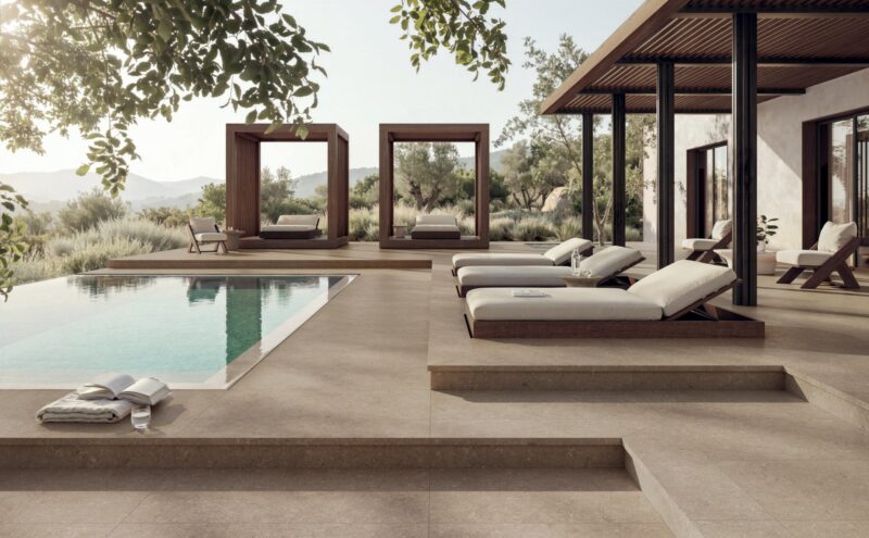 A poolside patio featuring lounge chairs, wooden cabanas, and views of a hilly landscape.