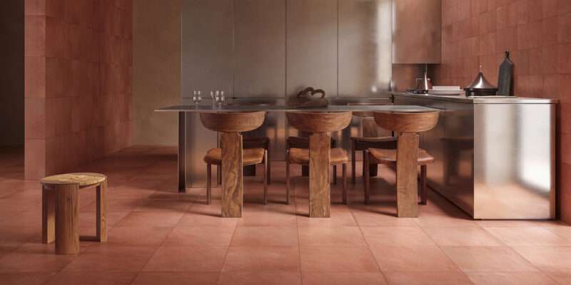A stainless steel kitchen island with three wooden chairs, a matching wooden stool in the foreground, and terracotta-colored floor and wall tiles.