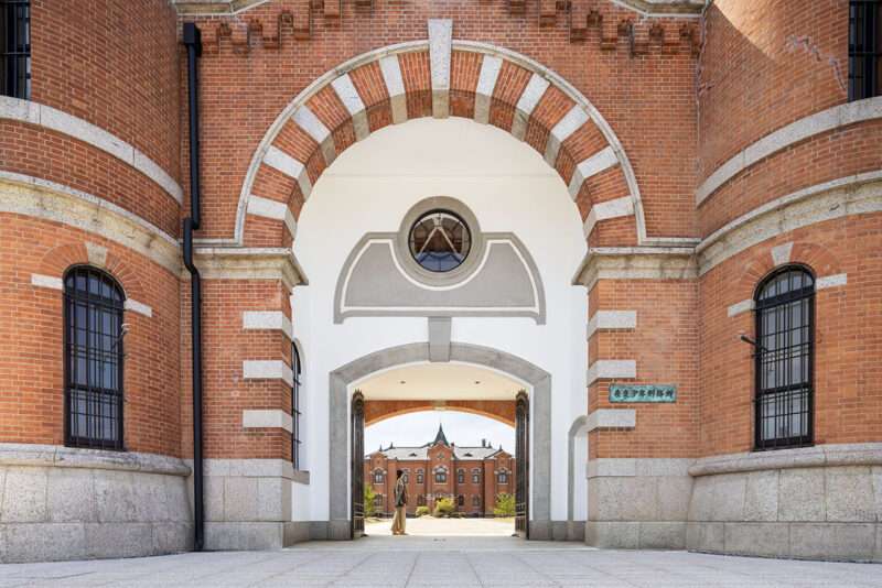 A person stands in the arched brick gateway of a historic building, with a matching brick building visible in the distance.