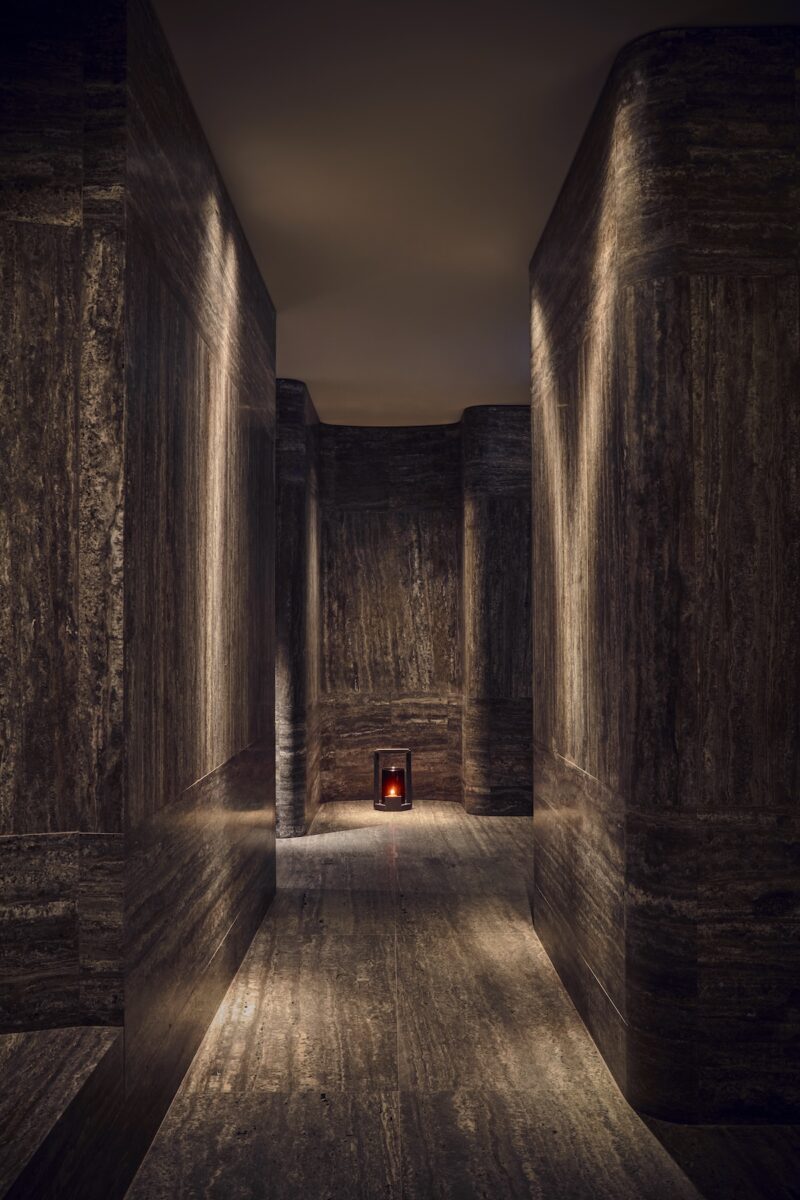 Dimly lit corridor with smooth, dark stone walls and floor, featuring a small candle lantern at the end of the hallway.