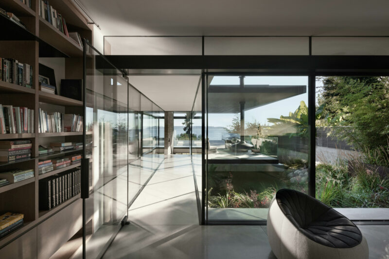 A West Coast modern home hallway with floor-to-ceiling bookshelves, glass walls, and views of a patio and the ocean.