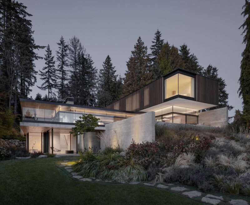 Modern multi-level home with floor-to-ceiling windows, built into a hillside surrounded by forest at dusk.