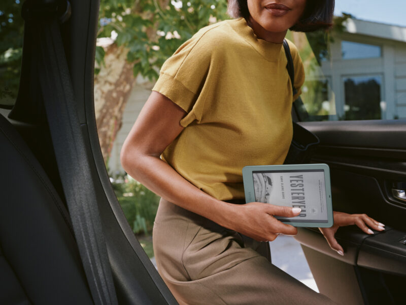 A woman in a mustard shirt exits a car while holding a Kindle redesign e-reader.