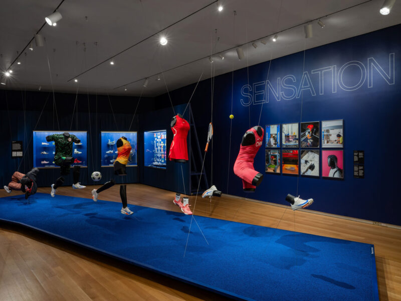 Museum exhibit Nike: Form Follows Motion, featuring floating athletic mannequins in sportswear, with framed photographs and the word “SENSATION” on the wall.