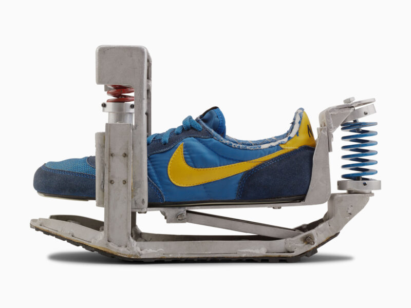 A blue and yellow Nike sneaker mounted in a metal mechanical frame with springs at the toe and heel, representing the concept of form follows motion.