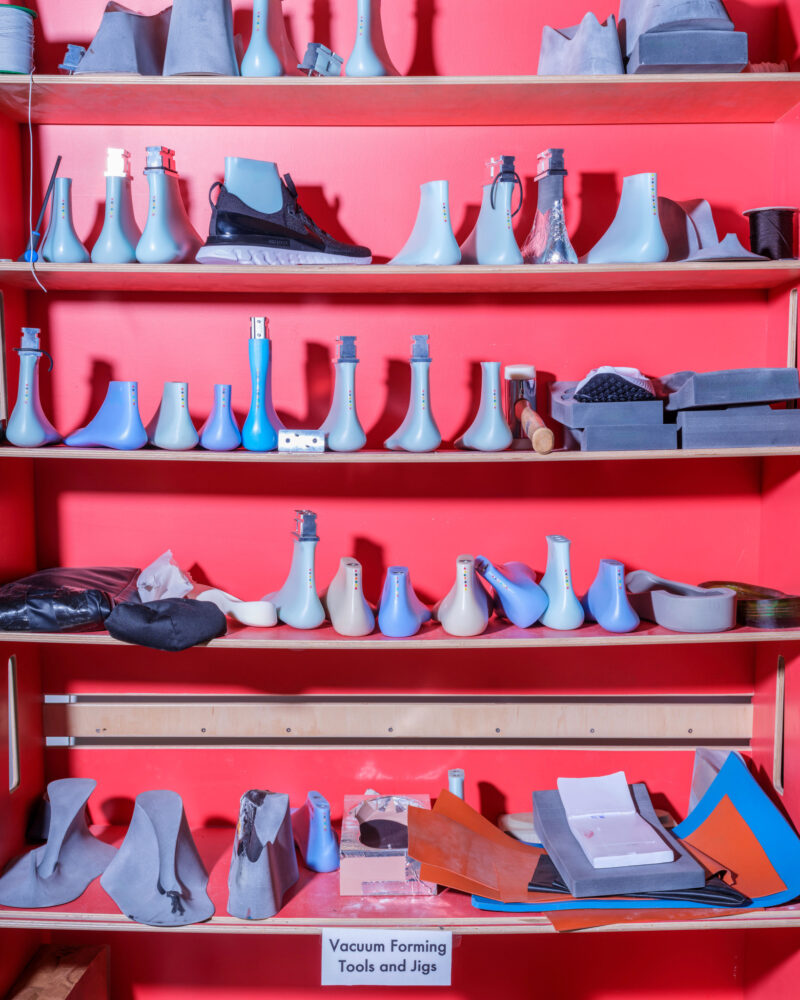 A red storage shelf holding rows of plastic shoe lasts and industrial molds, showcasing Nike: Form Follows Motion with a sign reading “Vacuum Forming Tools and Jigs” at the bottom.