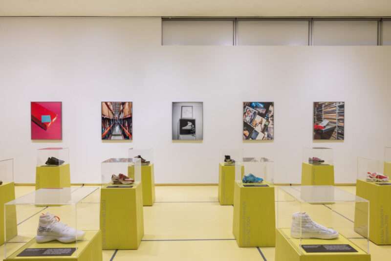 Sneakers from Nike: Form Follows Motion displayed on yellow pedestals under glass cases, with corresponding photographs hanging on the wall behind them.