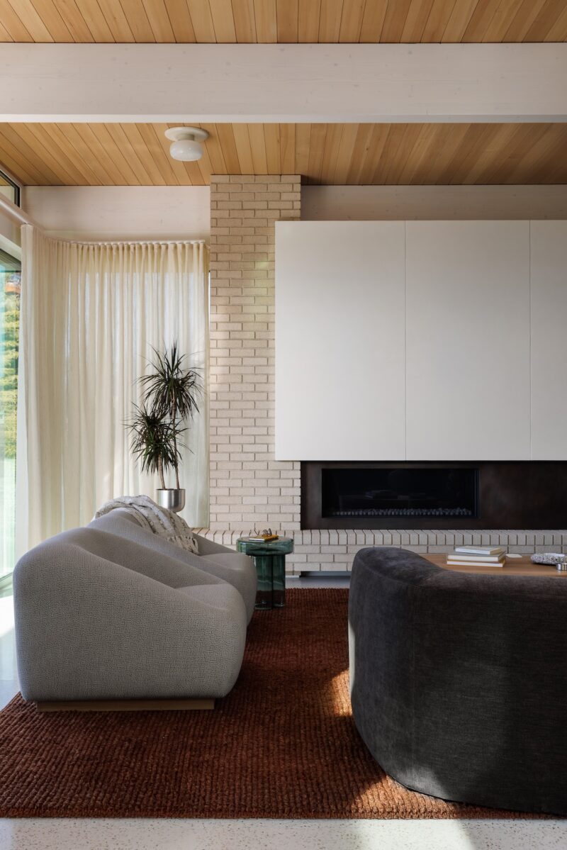 Modern living room with a light gray sofa, dark gray curved chair, brown rug, potted plant, and white brick fireplace with white cabinets above.