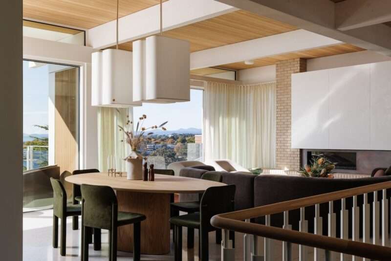 Modern dining and living area with wooden table, green chairs, pendant lights, large windows, neutral decor, and a view of trees and mountains outside.