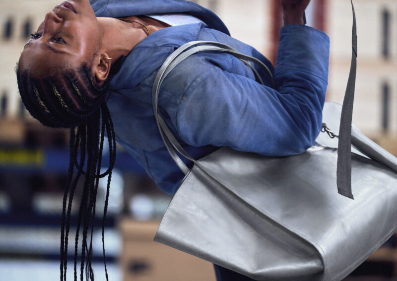 A person with braided hair wearing a blue jacket carries a large silver Freitag Reference collection tote bag.