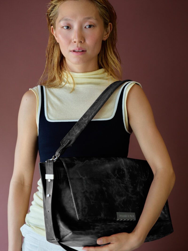 A person wearing a layered top carries a black messenger bag from the Freitag Reference collection over their shoulder.