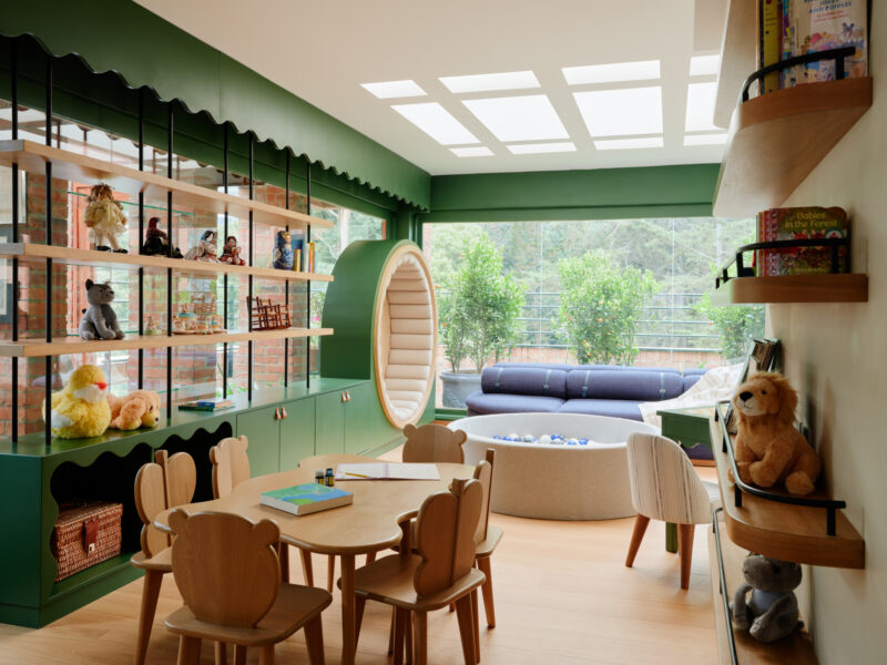 Children’s playroom with wooden tables and bear-shaped chairs, a circular reading nook, a ball pit, shelves filled with toys and books, and a view of trees through large windows.
