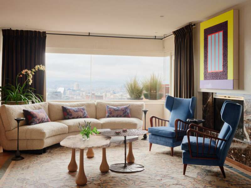 A living room with a curved, cream-colored sofa, two blue armchairs, and two tiered coffee tables in front of a window overlooking a city.