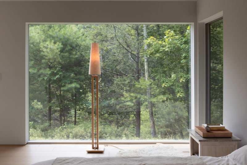 Tall wooden floor lamp with a conical shade in front of a large window overlooking a forest.