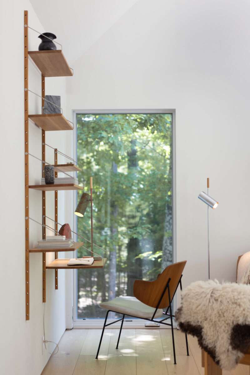 A modular wall-mounted shelving unit with a desk surface sits beside a vertical window and a wooden chair.