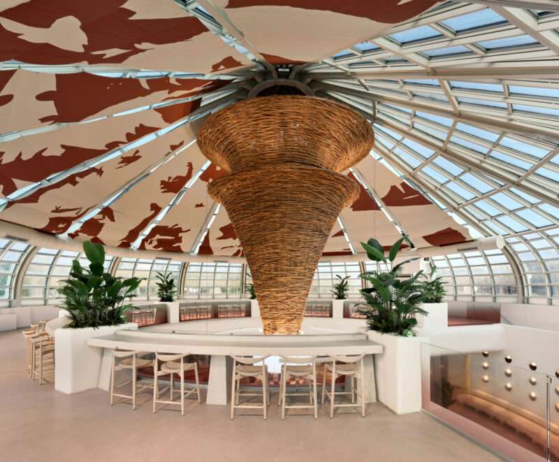 A curved bar with wooden stools beneath a large, woven, cone-shaped pendant light in a Local People greenhouse-style cafe with a glass-paneled domed ceiling.