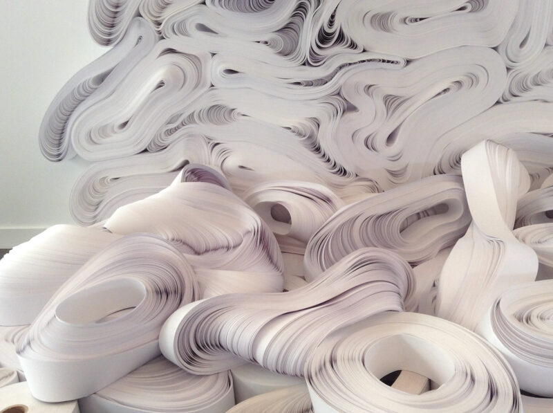 Large rolls and undulating strips of white paper stacked in a pile, evocative of Jae Ko sculptures.