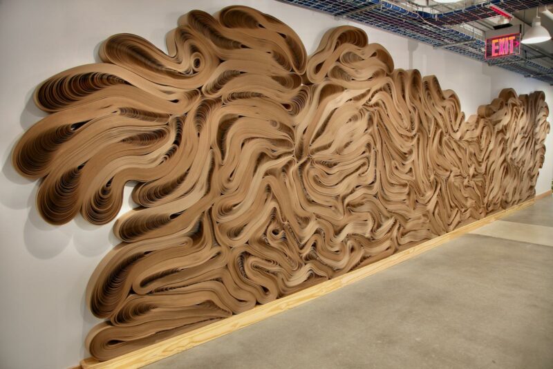 Large wall installation by Jae Ko featuring layered, swirling brown cardboard strips.