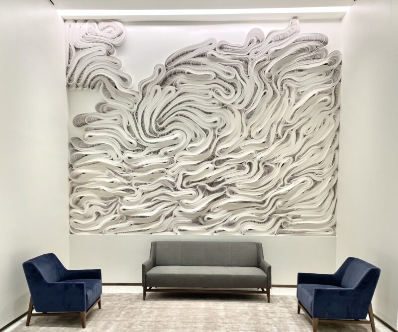 A gray sofa sits between two blue armchairs in front of a wall-mounted Jae Ko sculpture made of layered, swirling white paper.