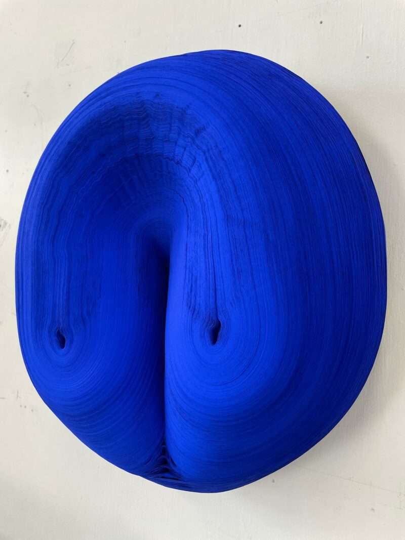 A circular, bright blue Jae Ko sculpture featuring a dense, layered texture with a central indentation.