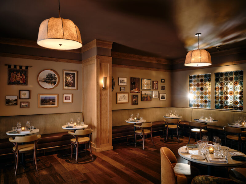 Restaurant interior with round tables, light-colored chairs, booth seating, and walls decorated with framed art and a circular glass bottle display.