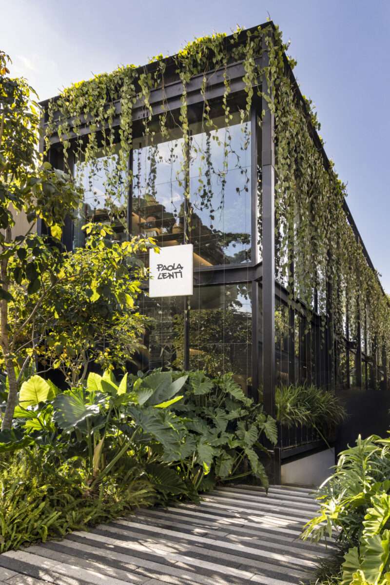Modern glass building with black frame and lush green vines cascading from the roof; a white sign reads “Paola Lenti.” Large leafy plants line the entrance steps on a sunny day.