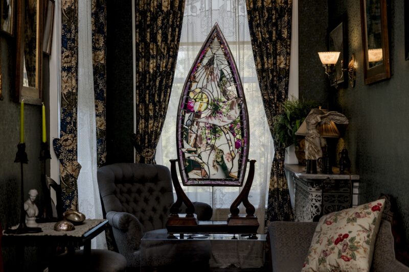 A stained-glass window in a pointed arch frame sits in broken light on a table between two upholstered chairs in a room with patterned curtains.