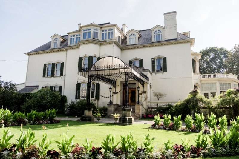 A three-story historic home with white brick siding and a decorative metal entryway awning stands in broken light, surrounded by landscaped gardens and a lawn.