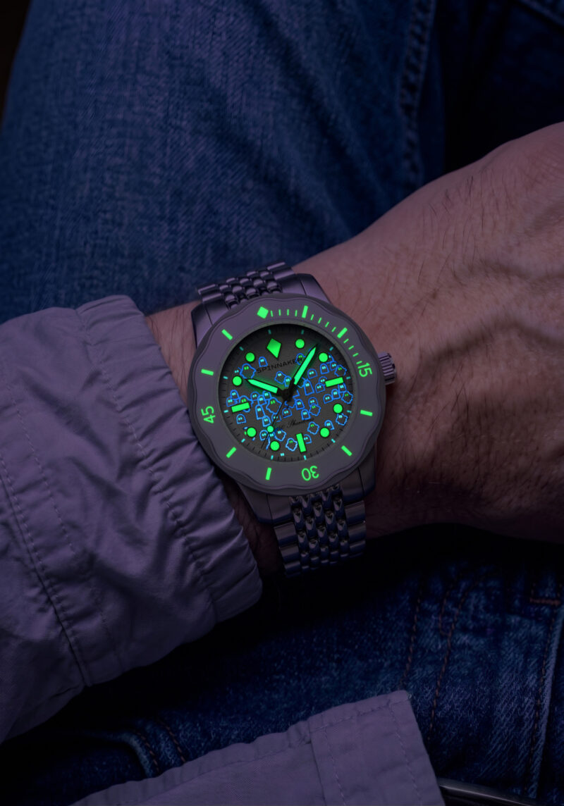 A Spinnaker 50 Phantoms dive watch on a person’s wrist, featuring a glowing luminous dial with a pattern of small diver helmet icons.