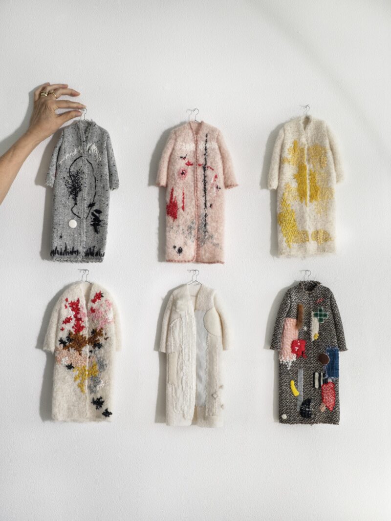 Six small, textured, patterned coat-shaped art pieces hang on a white wall; a hand adjusts the top left piece.